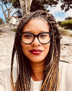 black woman with small locks and glasses. She is wearing a red lipstick with a tan cardigan. Her background looks like a beach setting with trees in the distance